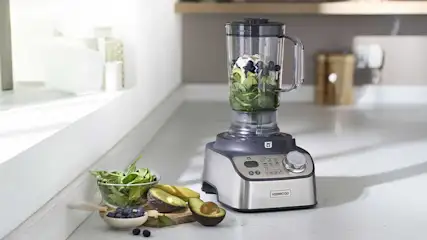 Een krachtige blender met metalen behuizing staat op een aanrecht; in de kan liggen spinazie, avocado, bosbessen en ijsblokjes voor een gezonde smoothie.