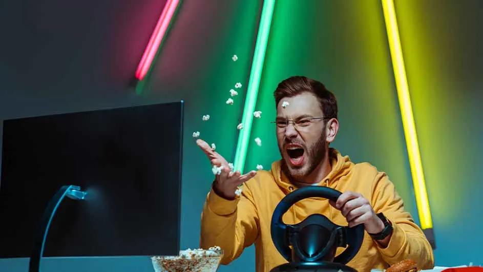 Een man met een geel shirt speelt een racespel met een stuurwiel, terwijl popcorn in de lucht vliegt voor een kleurrijke achtergrond.