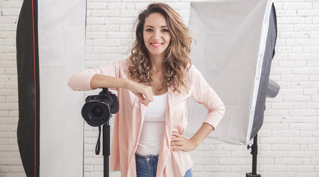 Een vrouw staat voor een camera op een statief, met een softbox en een bakstenen muur als achtergrond. Ze draagt een roze vest en blauwe jeans.