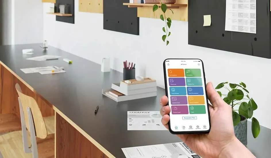 Hand houdt een smartphone met kleurrijke productiviteitsapp boven een lange werktafel met papieren, planten en bordjes.