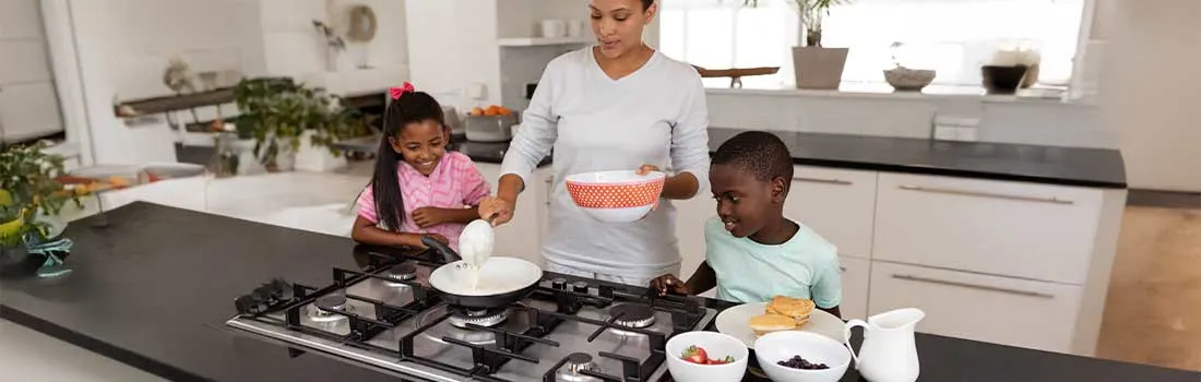 Moeder bakt pannenkoeken op het gasfornuis, terwijl haar twee lachende kinderen naast haar staan en samen genieten van het ontbijt.