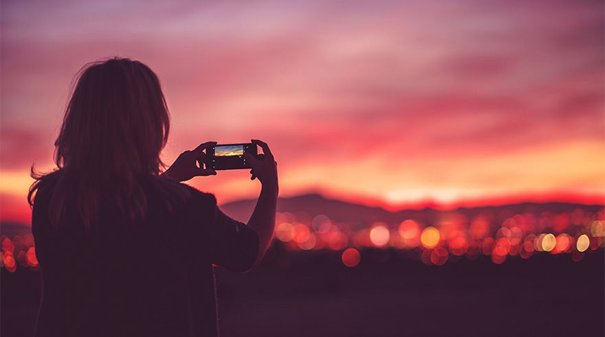 Een persoon fotografeert een zonsondergang met een telefoon. De lucht is roze en oranje, met lichten van een stad in de verte.