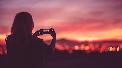 Een persoon fotografeert een zonsondergang met een telefoon. De lucht is roze en oranje, met lichten van een stad in de verte.