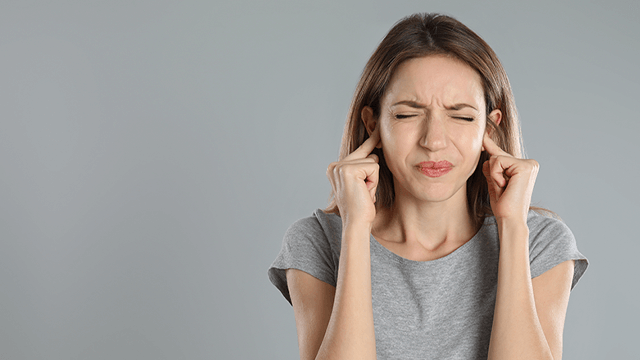 Een vrouw met een grijs t-shirt bedekt haar oren met haar vingers tegen een grijze achtergrond.