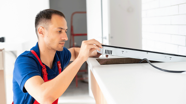 Een man in blauw shirt met rode schort installeert een wit fornuis tegen een witte muur met tegels in een keuken.