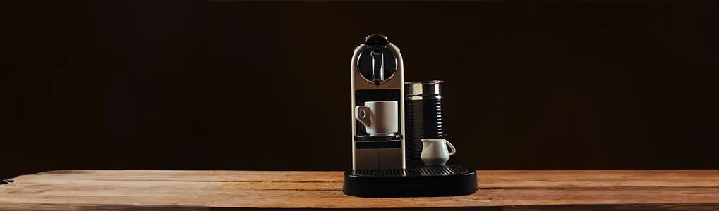 A coffee machine sits on a wooden table with a cup, frother and milk jug against a dark background.