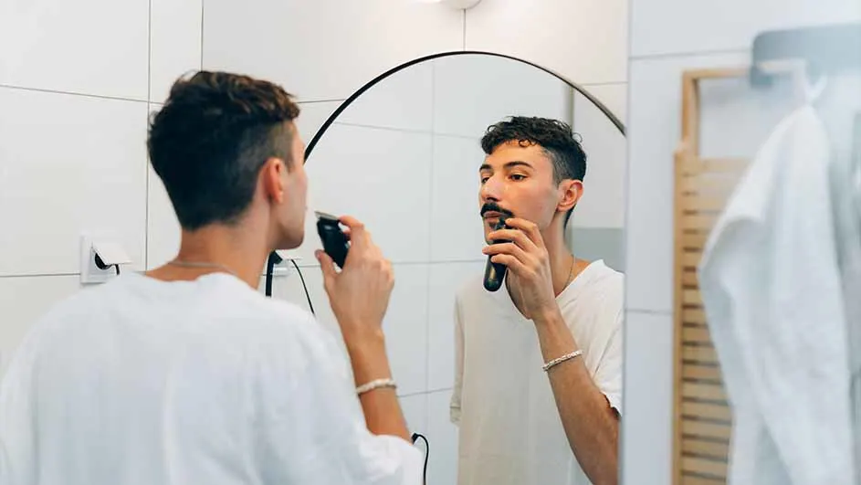 Een man in een witte top trimt zijn snor met een zwart elektrisch apparaat voor een ronde spiegel. De achtergrond toont witte tegels.