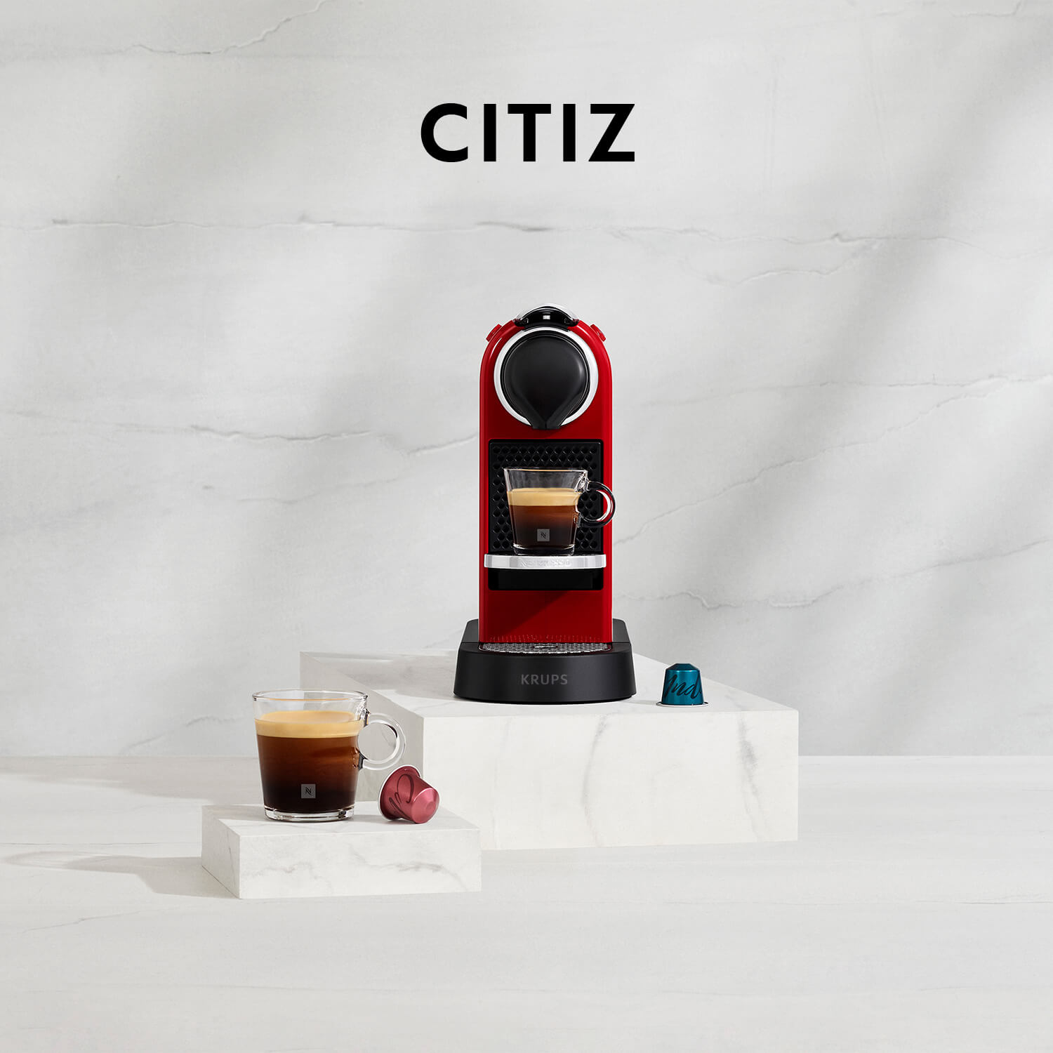 Rode Nespresso Citiz machine met kopjes espresso en capsules op witte oppervlakken, tegen een witte achtergrond.