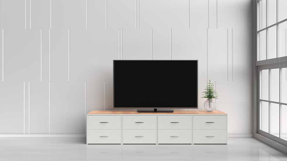 Een zwarte televisie staat op een witte kast met houten blad, voor een witte wand. Een raam staat rechts.