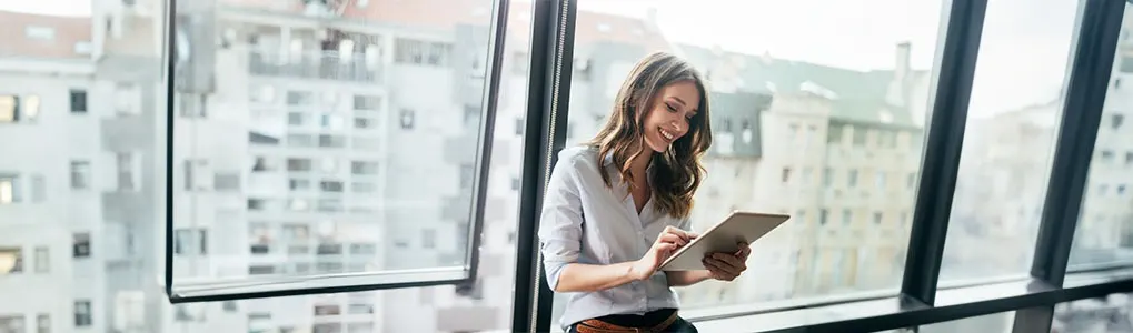 Vrouw gebruikt een tablet bij een groot raam. Op de achtergrond zijn gebouwen te zien.