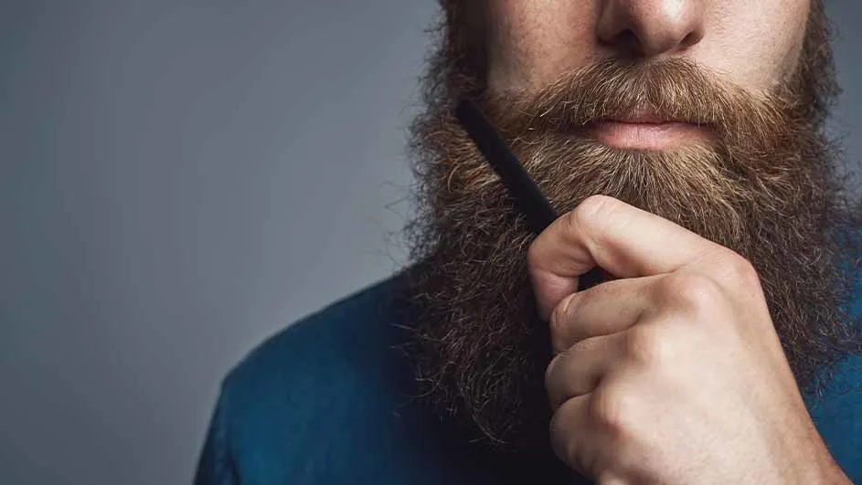 Een man met een baard kamt zijn baard met een zwarte kam tegen een grijze achtergrond.