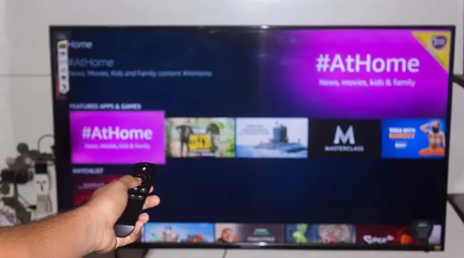 Een hand houdt een afstandsbediening voor een tv-scherm met een paarse #AtHome-interface en app-pictogrammen.