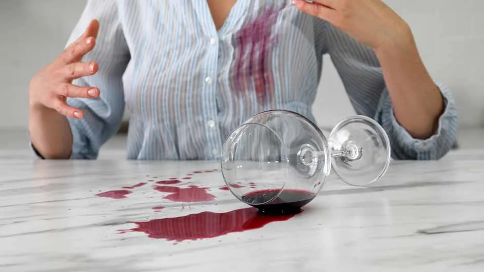 Een omgevallen wijnglas op een tafel met rode wijn, de wijn heeft ook een vlek op een blauw gestreept overhemd gemaakt.