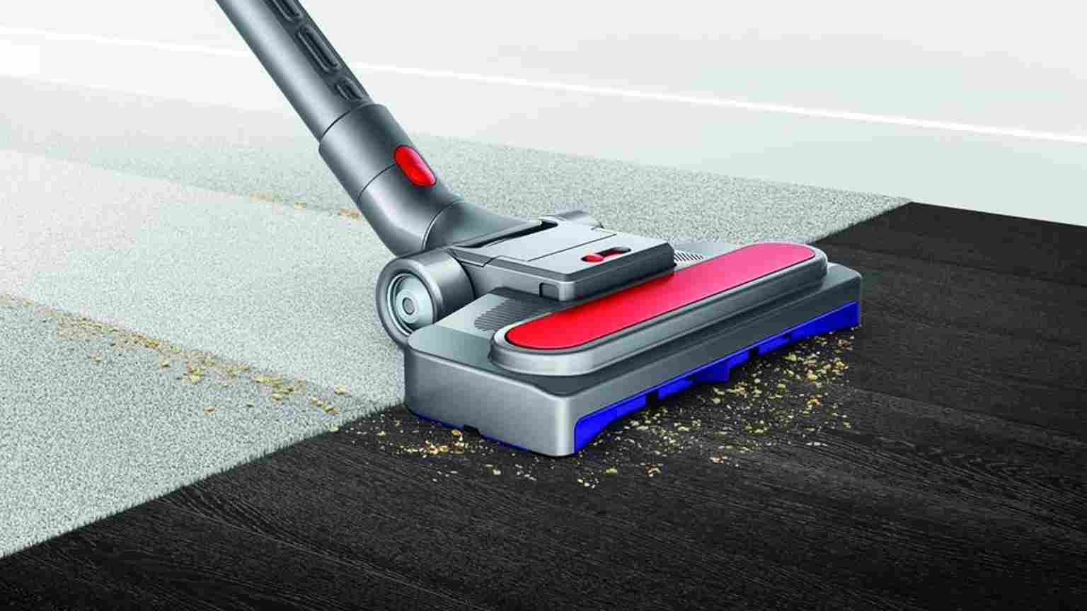 Een grijze stofzuiger met rode en blauwe accenten zuigt vuil op een vloer die half tapijt, half donker hout is.