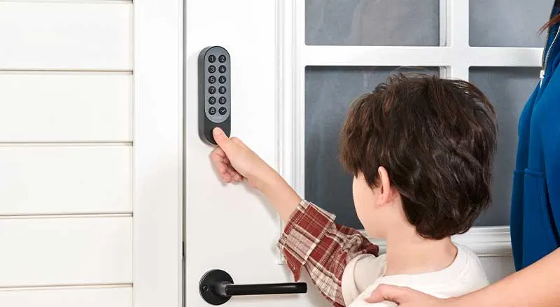 A child opens a door using a smart keypad, while an adult stands beside and watches.