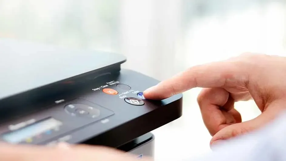 Vinger drukt op de blauwe kopieerknop van een zwarte all-in-one printer; bedieningspaneel met stop, zwart en kleur zichtbaar.