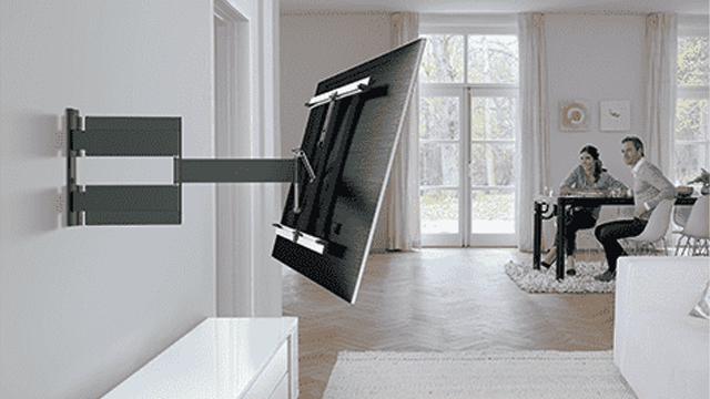 Een tv aan een grijze muurbeugel. Op de achtergrond zitten twee mensen aan een tafel bij het raam.