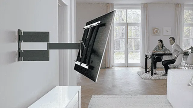 Een tv aan een grijze muurbeugel. Op de achtergrond zitten twee mensen aan een tafel bij het raam.