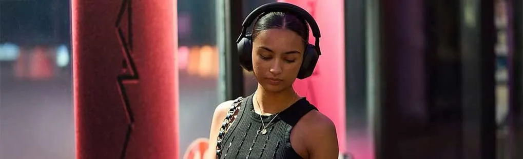 Een vrouw met zwarte draadloze Sony koptelefoon loopt buiten in een zwarte top, met een ketting en een tas over haar schouder.