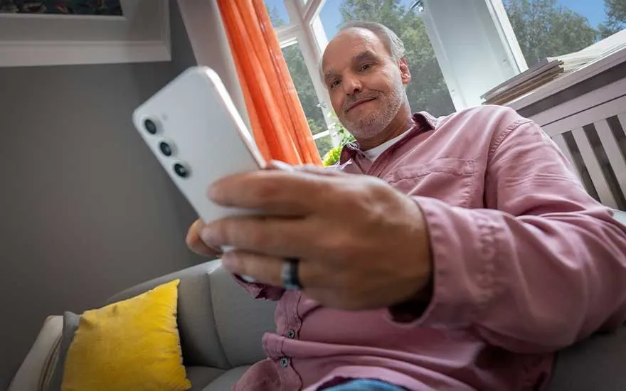 Man sits comfortably on his sofa at home, casually browsing his modern smartphone near a bright window.