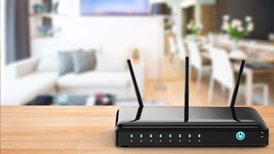 Een zwarte router met drie antennes op een houten tafel, voor een vage woonkamer met bank en eettafel.