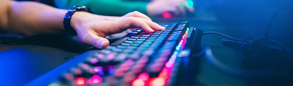 A person's hand presses keys on a backlit keyboard. A wrist watch is visible. A computer mouse sits on a desk.