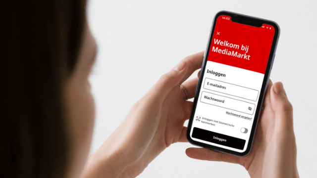 Persoon houdt smartphone vast met inlogscherm van MediaMarkt app, met rode header en inlogvelden zichtbaar.