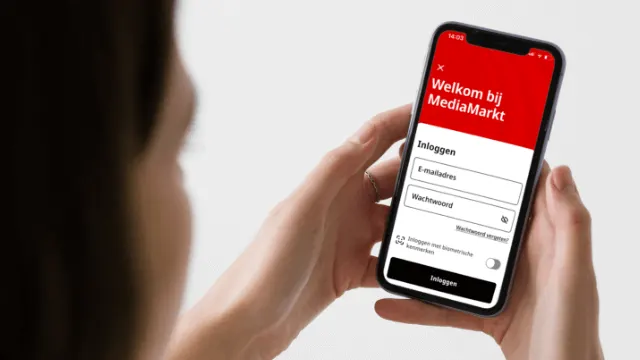 A person holds a smartphone with the MediaMarkt app login screen, with the red header and login fields visible.