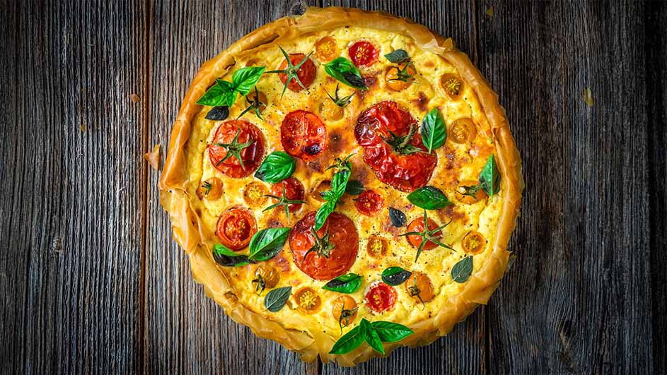 Een quiche met rode en gele tomaten en basilicum op een houten tafel.
