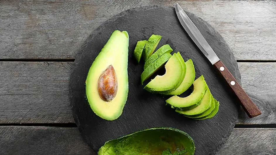 Een gehalveerde avocado met pit, schijfjes avocado en een mes op een zwart bord, tegen een houten achtergrond.