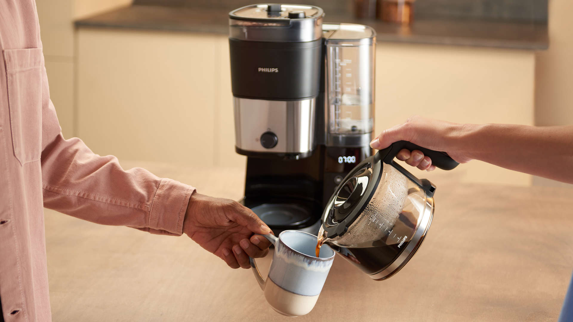 Koffie wordt gegoten uit een Philips koffiekan in een mok, naast een Philips koffiezetapparaat op een aanrecht.