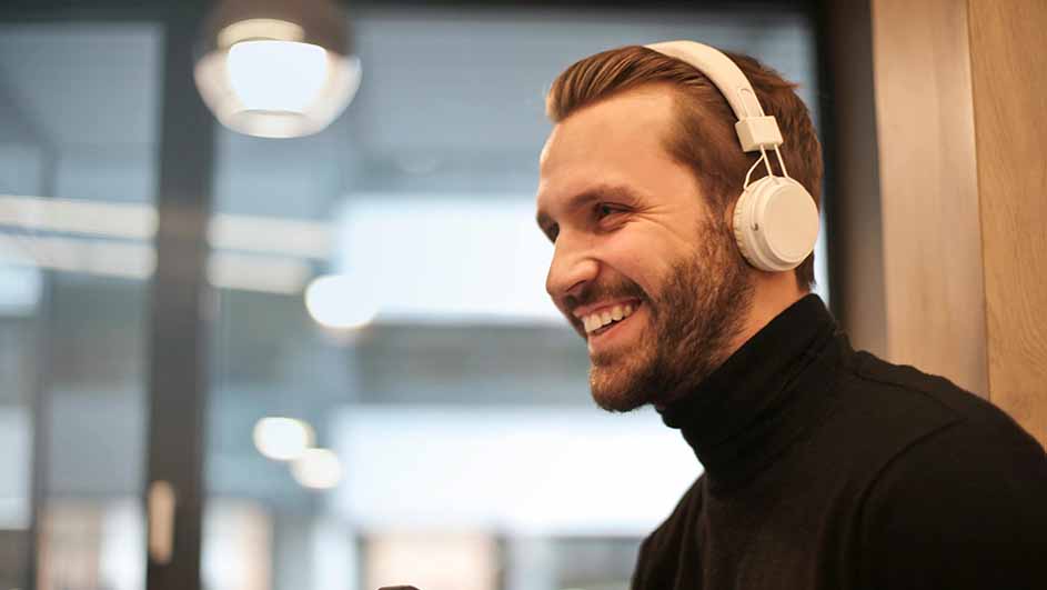 Een lachende man met een zwarte coltrui en witte hoofdtelefoon staat voor een wazig raam en een lamp.