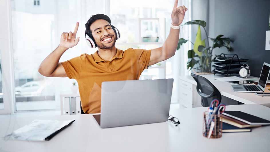 Man met koptelefoon danst achter een laptop op een wit bureau met kantoorartikelen, voor een raam en een plant.
