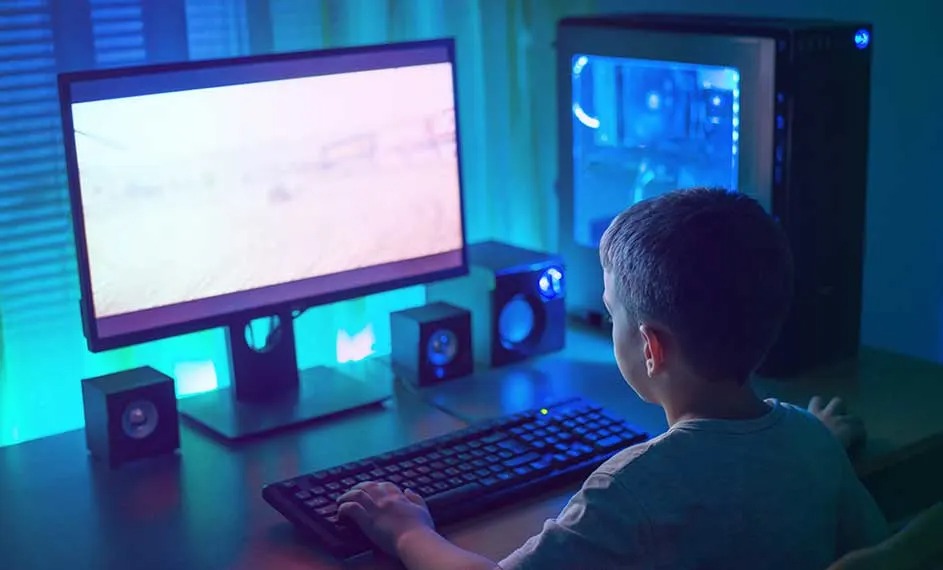Op de afbeelding zit een jongen achter een verlichte gaming-pc met toetsenbord, muis, speakers en monitor, in een kamer met blauwe sfeerverlichting.