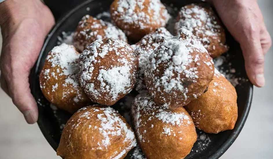 Een schaal vol versgebakken oliebollen bestrooid met poedersuiker wordt vastgehouden door twee handen. Het merk is niet zichtbaar.