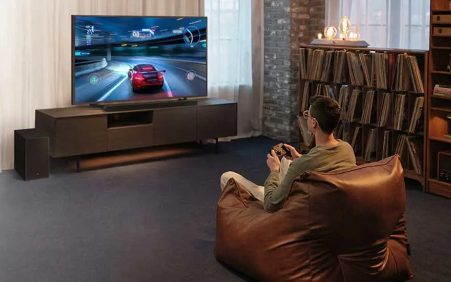 Man speelt racegame op groot scherm, zittend in zitzak, met soundbar, subwoofer en platenkast ernaast.