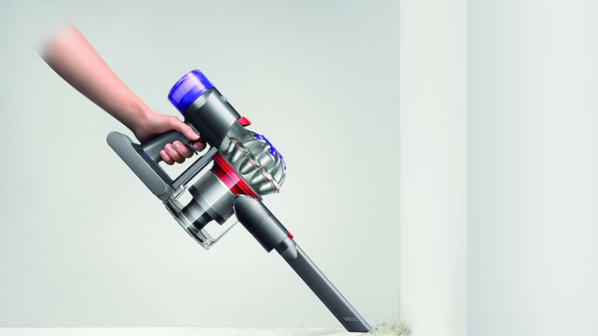Een hand houdt een grijze Dyson kruimeldief met blauwe en rode accenten vast, gericht op een plint met stof.