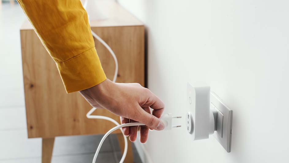 Een hand steekt een witte stekker in een stopcontact op een witte muur, met een houten kast op de achtergrond.