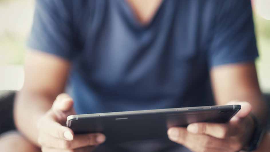 Een persoon met een blauw shirt houdt een zwarte tablet vast, met beide handen zichtbaar, tegen een vage achtergrond.