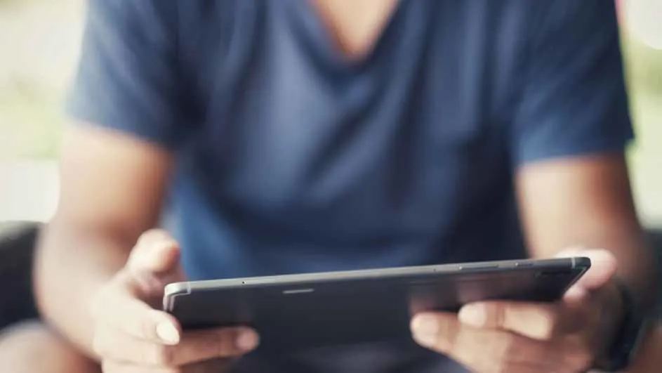 Een persoon met een blauw shirt houdt een zwarte tablet vast, met beide handen zichtbaar, tegen een vage achtergrond.