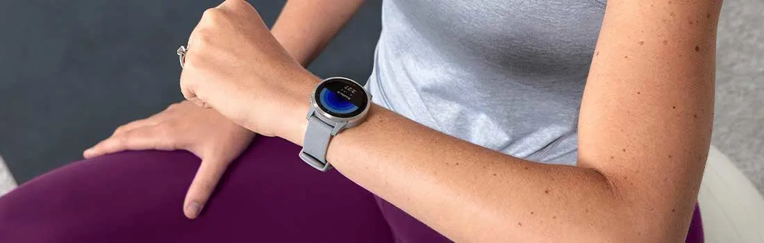 Een vrouw met een grijze sporttop en paarse legging draagt een grijze Garmin smartwatch, zittend op een fitnessbal of bank.