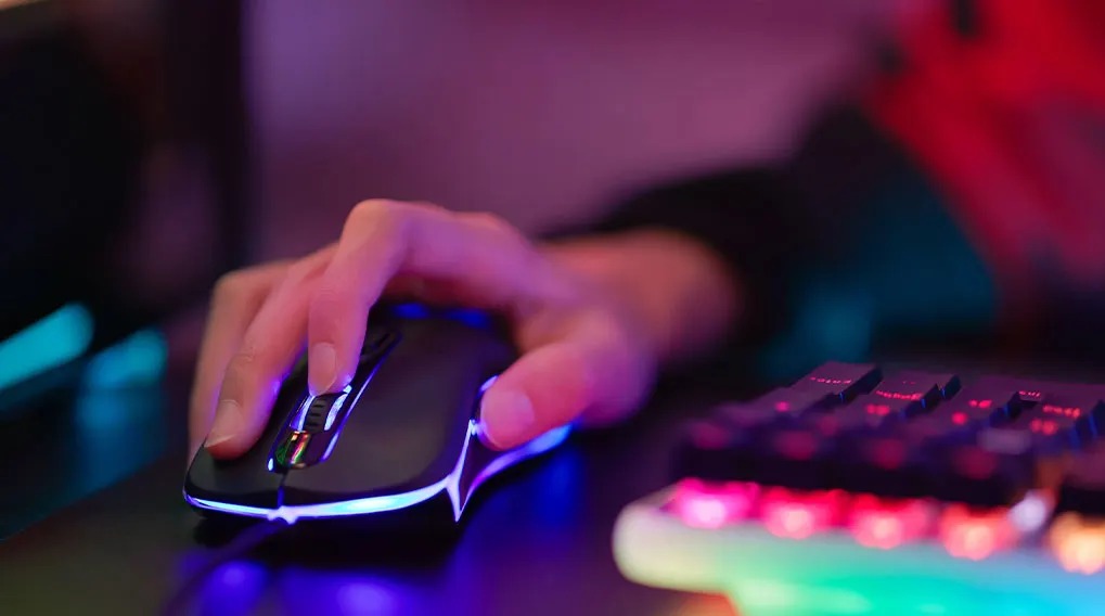 A hand uses a black computer mouse with blue lights. A keyboard with red and white lights is on a dark desk.