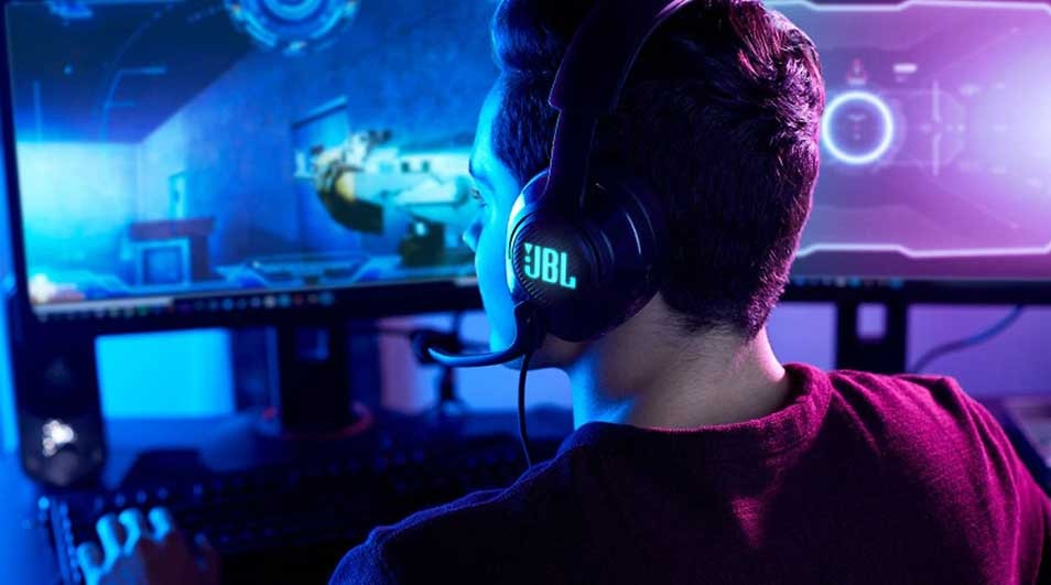 A focused gamer wearing a glowing JBL headset sits in cool blue light, facing an ultrawide monitor showing a futuristic game.