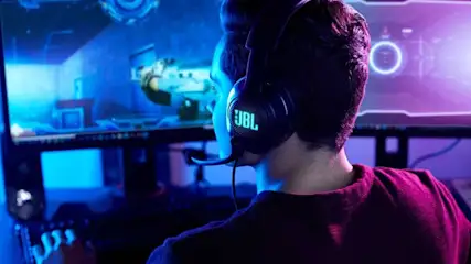 A focused gamer wearing a glowing JBL headset sits in cool blue light, facing an ultrawide monitor showing a futuristic game.