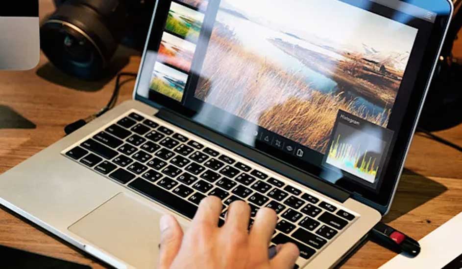 Op een houten bureau staat een laptop met een foto en histogram op het scherm, een hand typt, een camera staat links.
