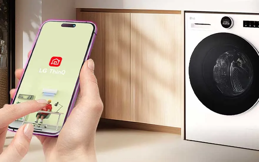 Hand opent de LG ThinQ-app op een smartphone naast een ingebouwde wasmachine in een warme, moderne wasruimte.