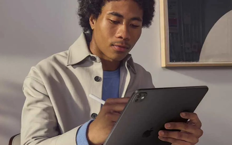Man in licht jack schrijft nauwkeurig met een stylus op een donkere tablet aan een tafel in een modern, minimalistisch interieur.