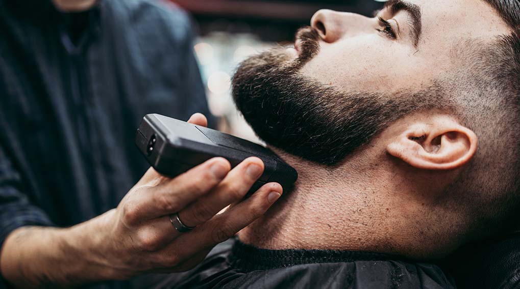 Een man krijgt zijn baard bijgewerkt met een zwarte trimmer door een kapper in een kapsalon.