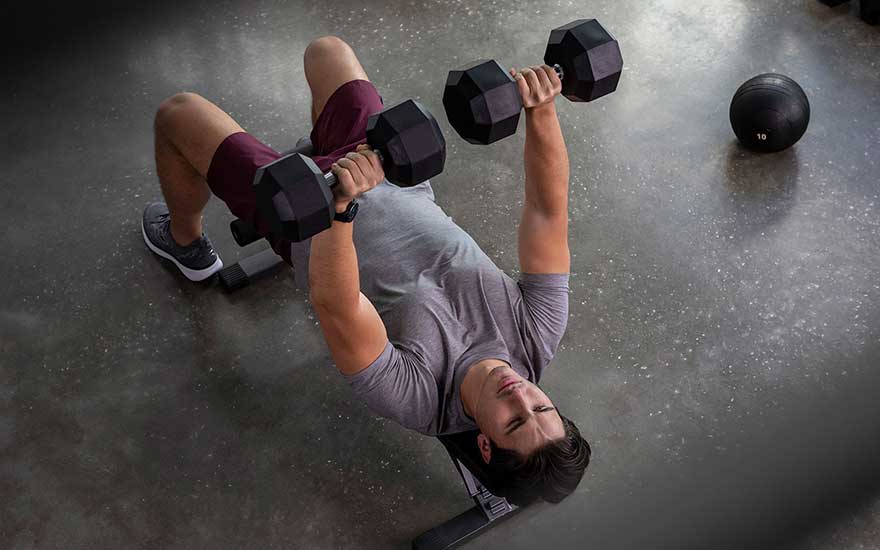 Man voert bankdrukken uit met twee halters, dragend een sporthorloge, op een bankje in een fitnessruimte.