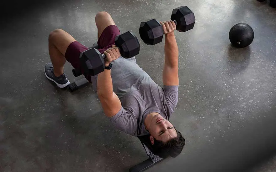 Man voert bankdrukken uit met twee halters, dragend een sporthorloge, op een bankje in een fitnessruimte.